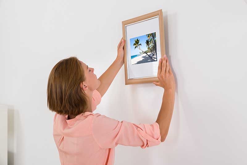 Young Woman Putting Photo Frame On White Wall_Andrey