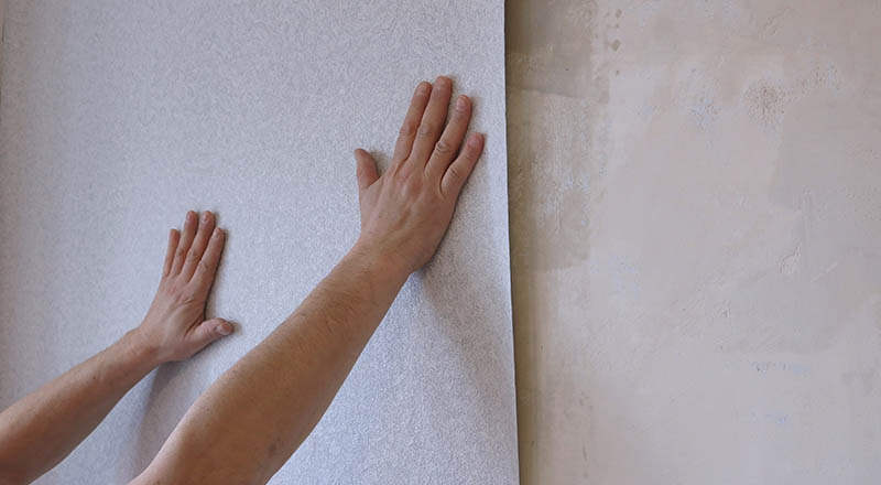 Hands smoothing a strip of light non-woven wallpaper on the wall